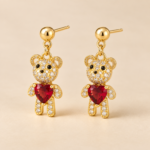 Gold-plated teddy bear drop stud earrings with clear CZ details and red heart-shaped feature stones, shown at an angle on a warm beige background.