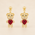 Pair of gold-plated teddy bear drop stud earrings with clear CZ detailing and red heart-shaped feature stones on a warm beige background.