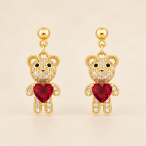 Pair of gold-plated teddy bear drop stud earrings with clear CZ detailing and red heart-shaped feature stones on a warm beige background.