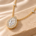 Close-up of a 9ct yellow gold pear lab-grown diamond halo necklace resting on warm beige stone, showing the setting and diamond-set chain stations
